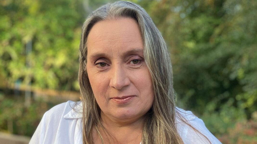 NHS trust to review 4,500 breast cancer cases after unnecessary surgeries found Denise Howarth. She has long fair hair and is wearing a white shirt. Behind her are some trees.