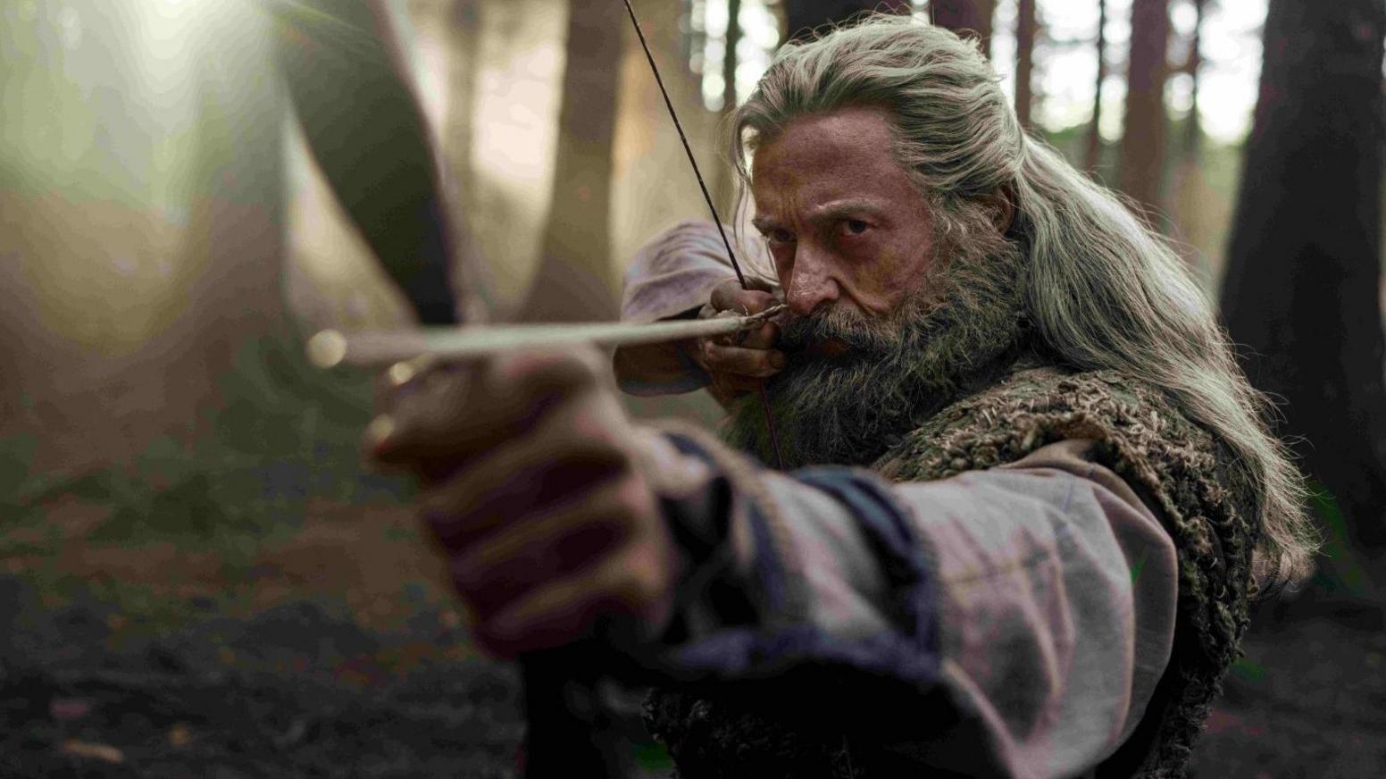 Hugh Jackman as Robin Hood. He has a large beard and grey flowing hair. He has a bow and arrow and the arrow is right at the end of the camera lens. He is standing in a wood.