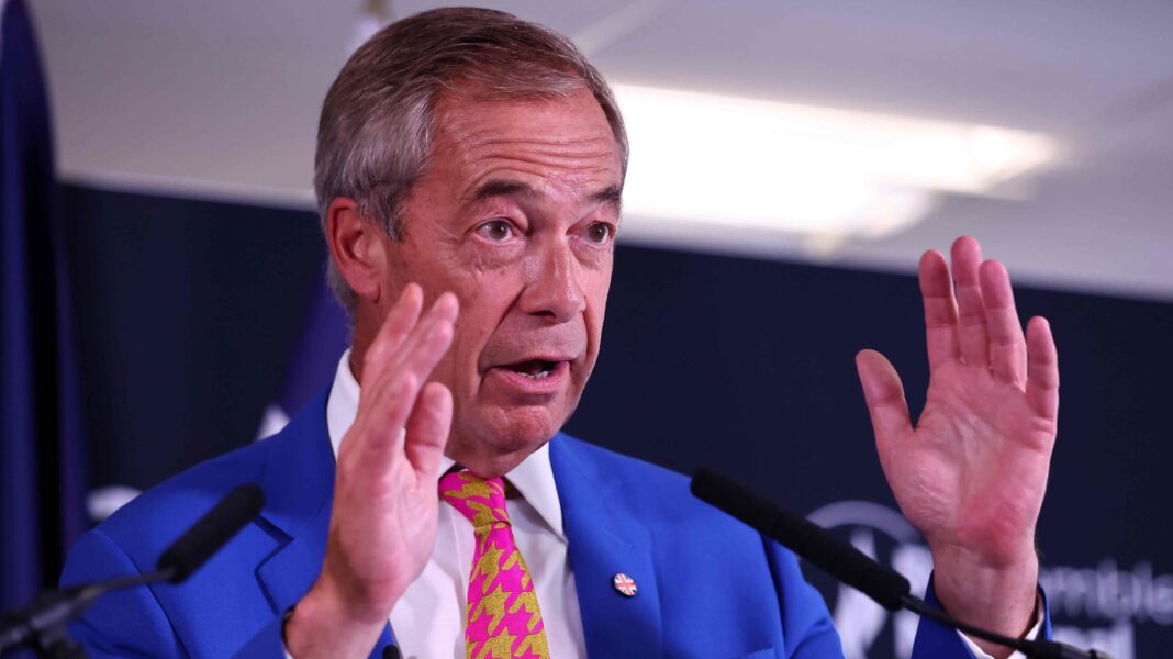 Nigel Farage speaking in front of two microphones, wearing a blue suit and a pink and yellow tie. He is wearing a union flag-patterned badge on his lapel.