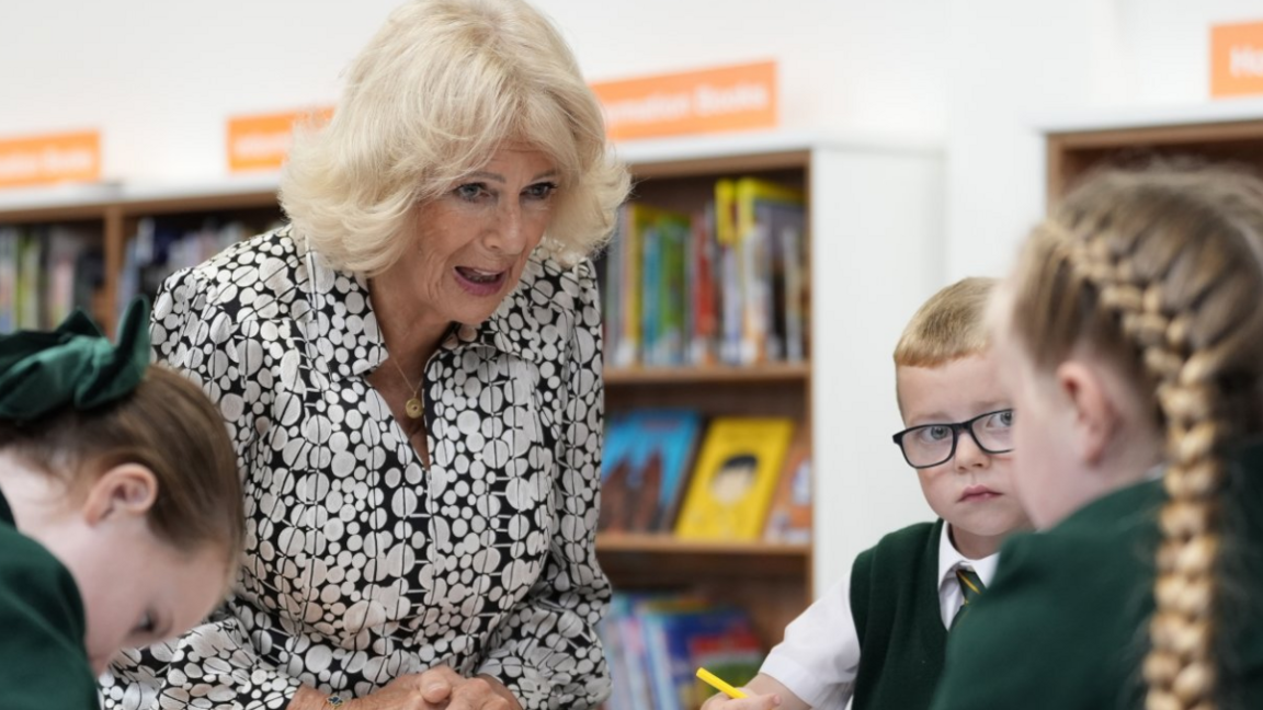 Queen Camilla in a dress of white circles on a black background talks to a boyu and two girls in green school uniform.
