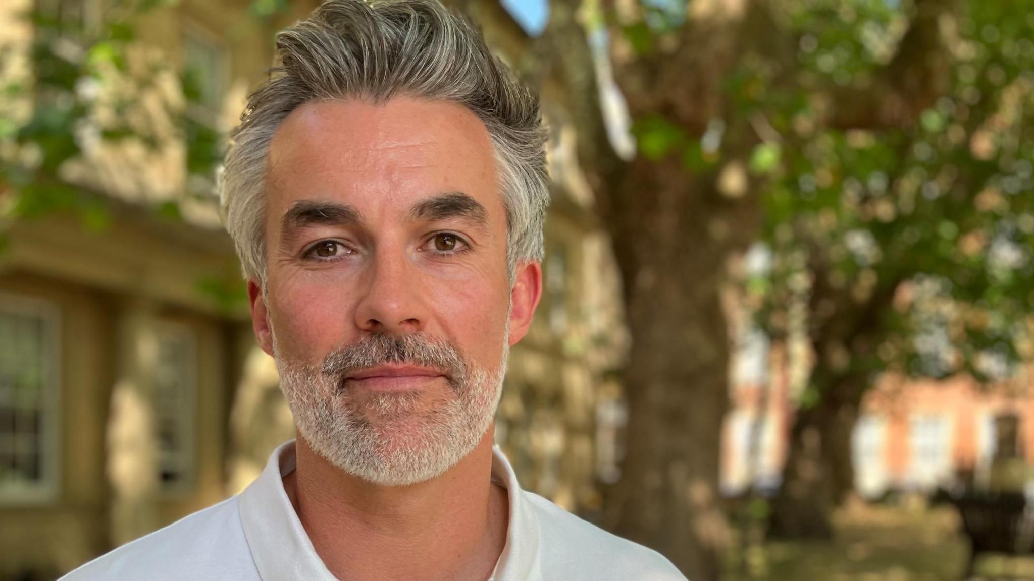 A man with greying hair and beard looks at the camera. In the background is a tree and red-brick building out of focus.