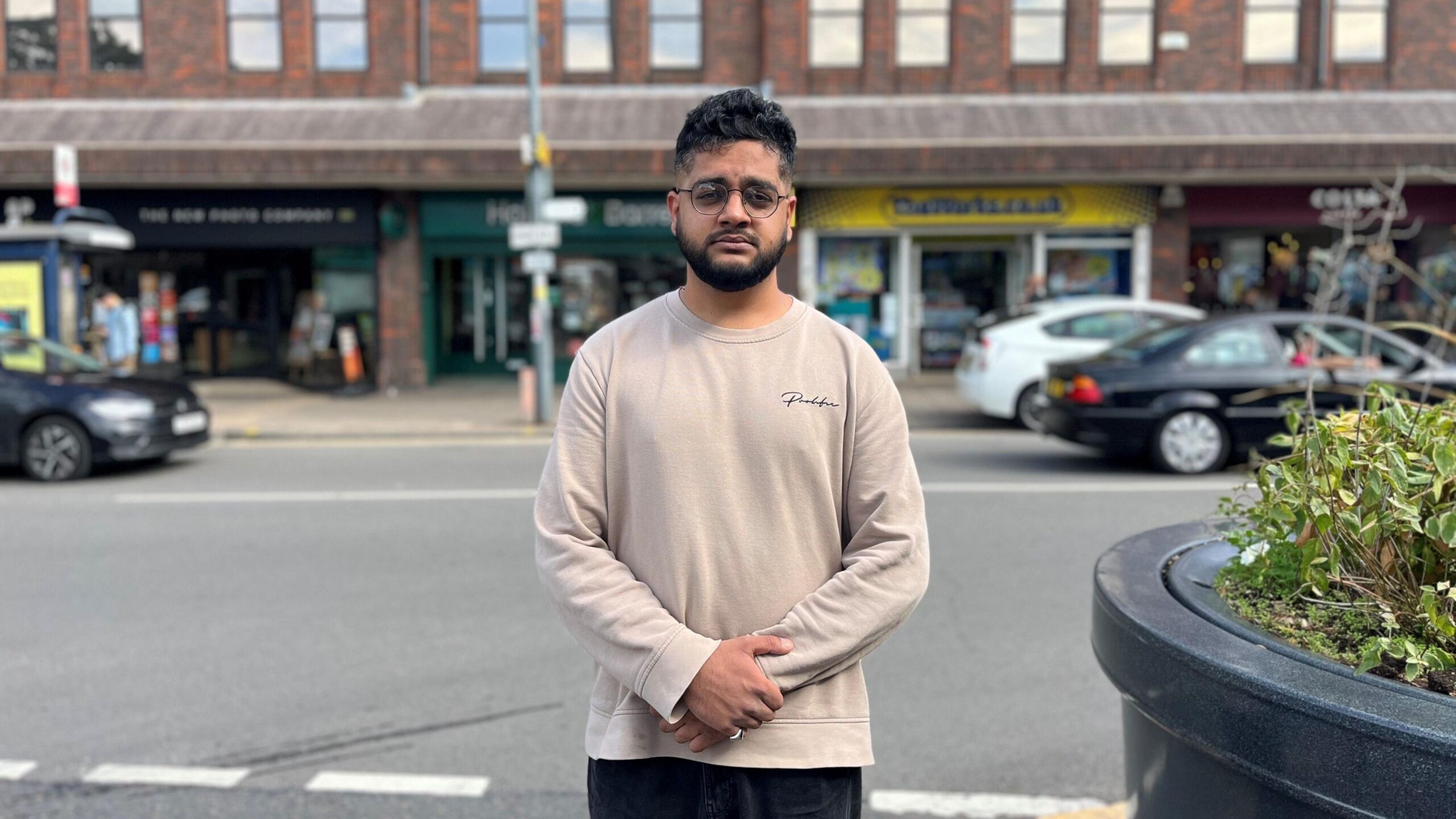 A man with dark short wavy hair, glasses and a moustache and beard is standing in front of a row of shops, which are blurred. He is wearing a cream long sleeved top and black trousers and his hands are linked in front of him.