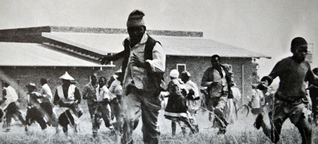A grainy black & white image of a group of people running towards the camera. A low-rise building can be seen behind them.