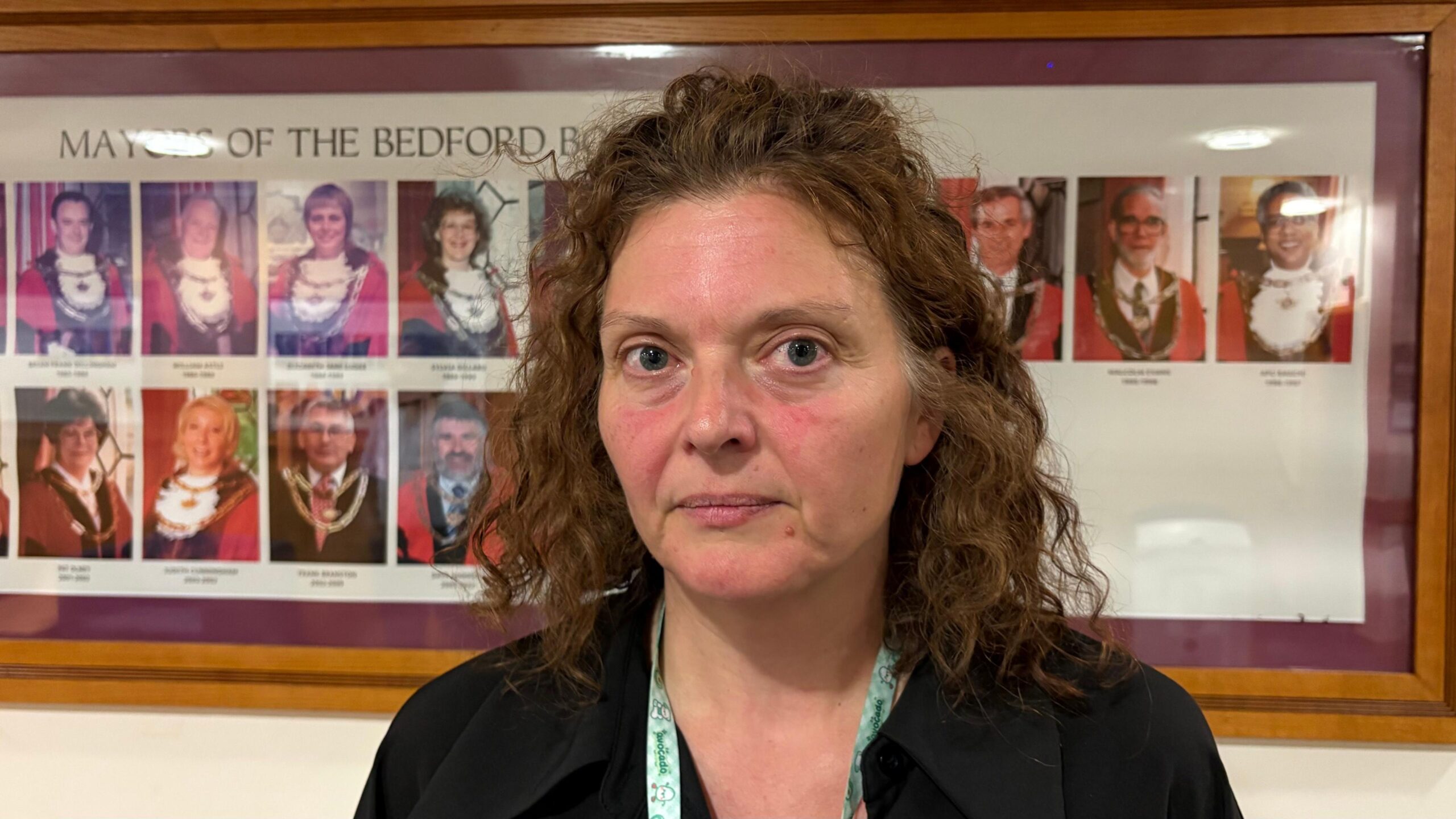 Zara Layne with curly hair stands in front of a framed display showing photographs of former mayors. The display, mounted on a wall with a wooden frame, is headed “Mayors of the Bedford Borough” and contains rows of portrait photographs.  Councillor Layne is wearing a dark top and a lanyard, and is positioned in the foreground with the mayoral display visible behind. The image is taken indoors under bright lighting.