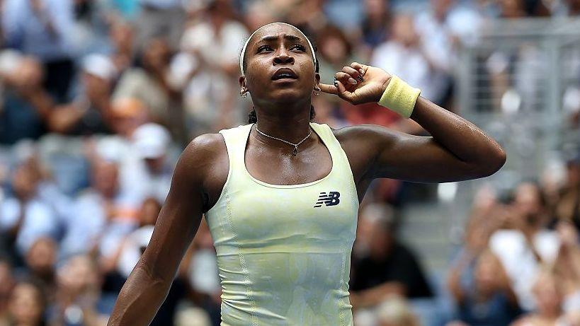 Coco Gauff, wearing a pale yellow vest, puts her left index finger behind her ear