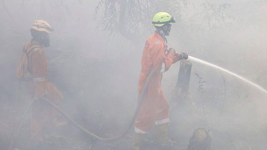 Two firefighters in bright orange uniform and helmets, standing outdoors in thick white haze, spray water from a hose