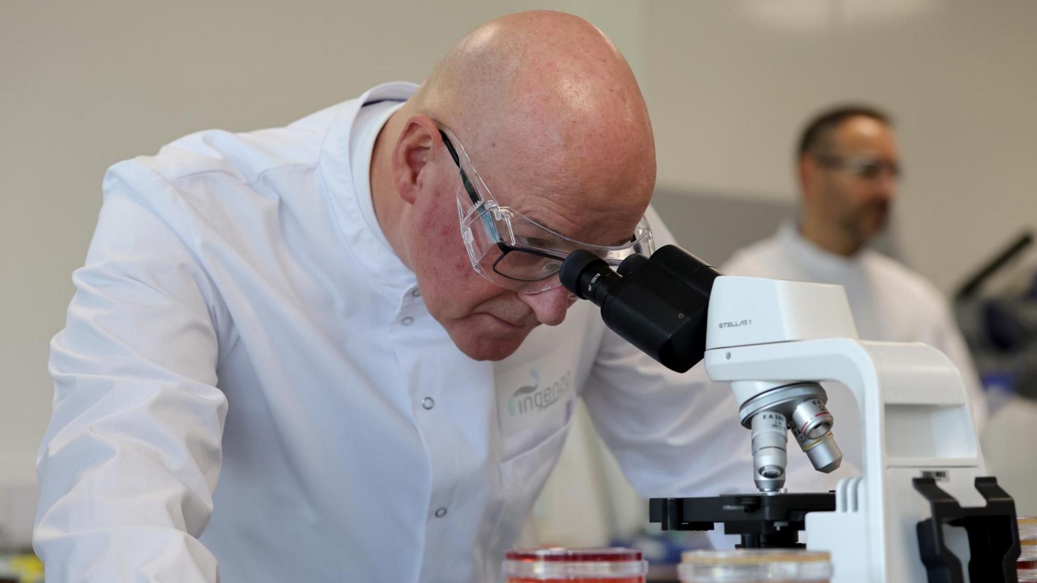 John Swinney, who has a bald head, is wearing transparent goggles over his dark-rimmed glasses. He is wearing a white lab coat and looking into a microscope.