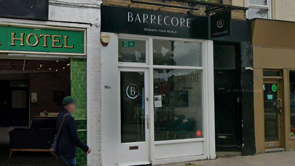 A white shop front on a high street with a black sign with the word Barrecore spelled out in a white font