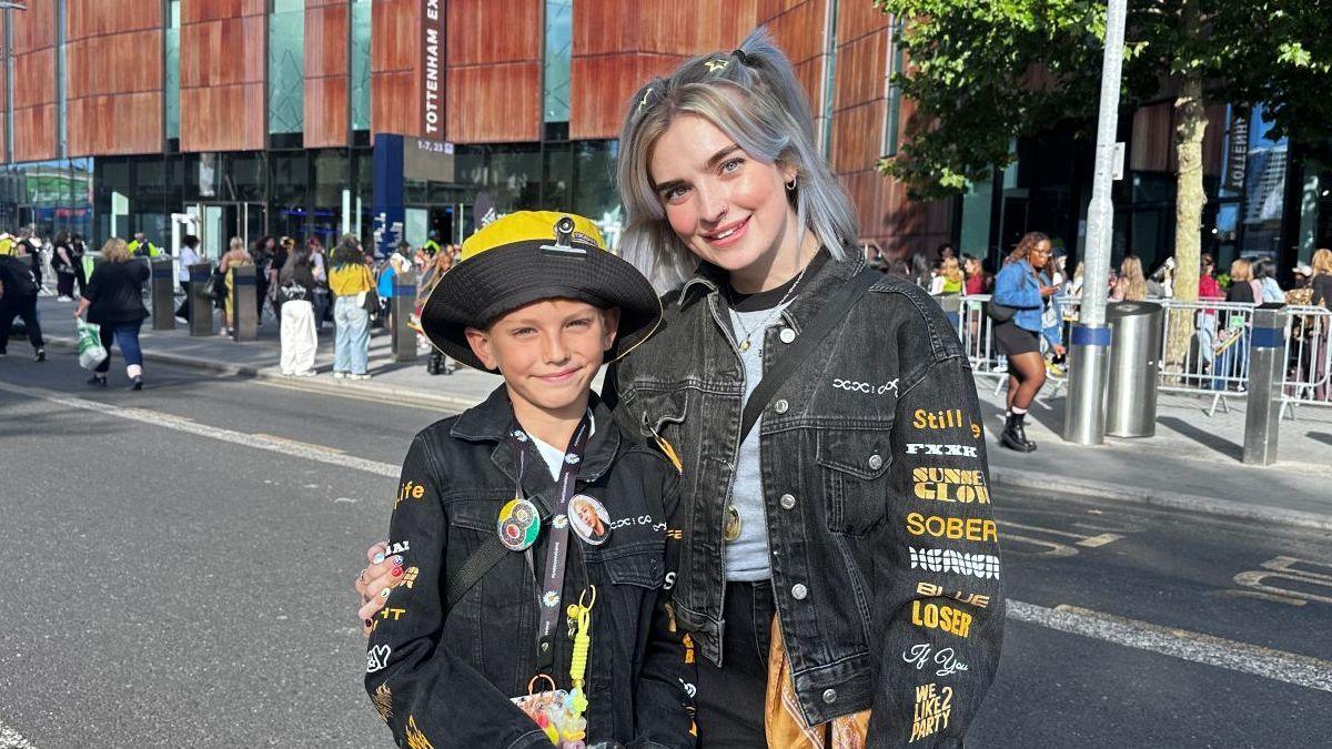 Mum and son standing in the foreground on a road outside a stadium. Both are wearing black denim jackets decorated with printed text, badges and pins. In the background, queues of people stand behind metal barriers outside the stadium entrance, with trees, bollards and signage visible along the pavement.