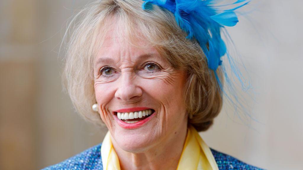 Esther Rantzen appears in a recent picture. She is smiling and dressed in a blue jacket with a matching feather in her hair.