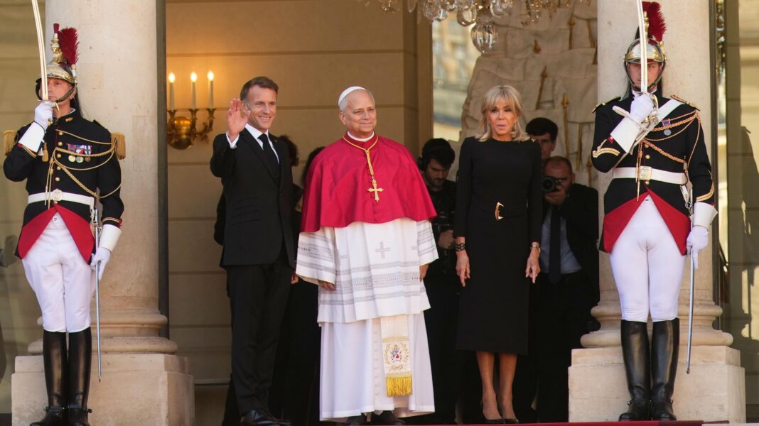 Hundreds of thousands expected in Paris for Pope’s visit Pope in centre, with a waving Macron on his right and Brigitte Macron on his left. There is a French guard standing on either end of the picture.