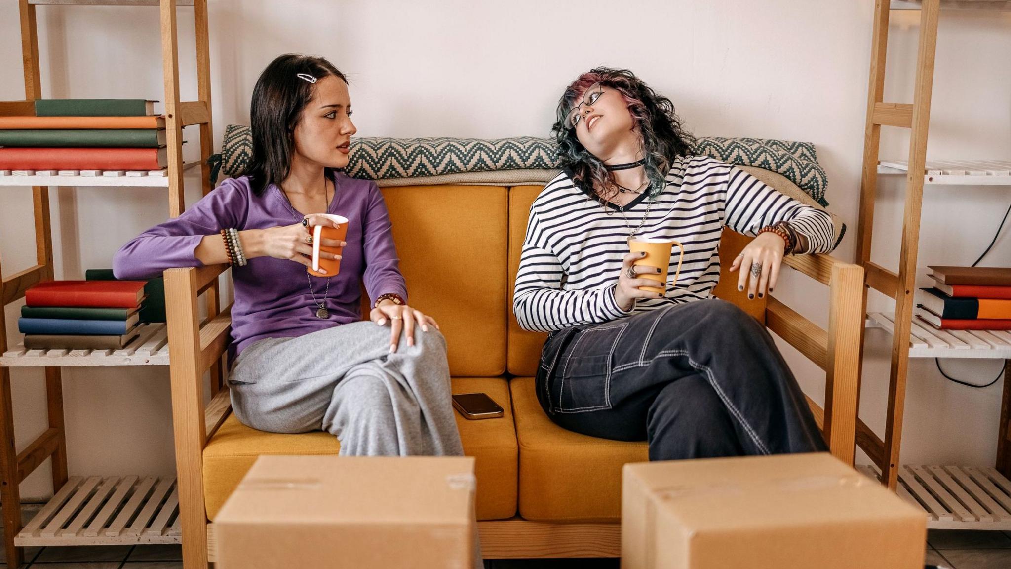 Two young women holding mugs sit on sofa surrounded by boxes and empty shelves.