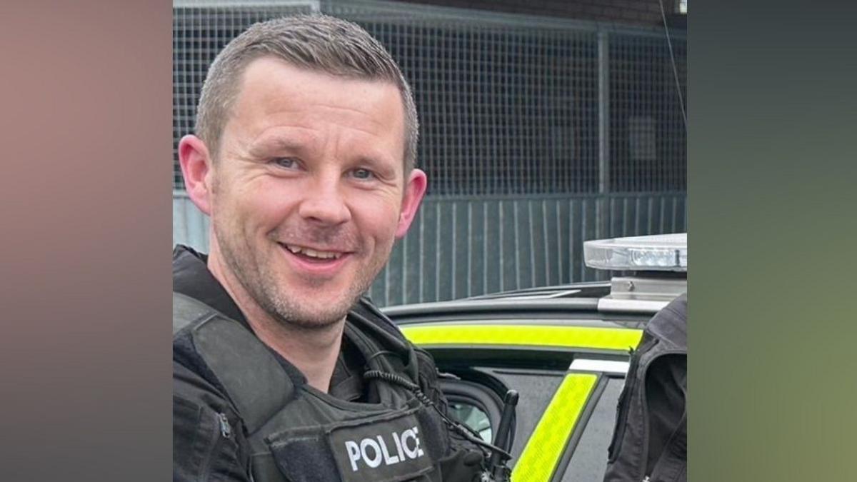 Tom Clough smiling into the camera. He has short, brown hair which is slightly longer on the top and styled to the left. His shoulders and chest are visible and he is wearing a black uniform with a bulky vest which has 'police' written on it in white letters. He is standing in front of a police car which is bright yellow with lights on the roof. There is a grey metal wall in front of the car.