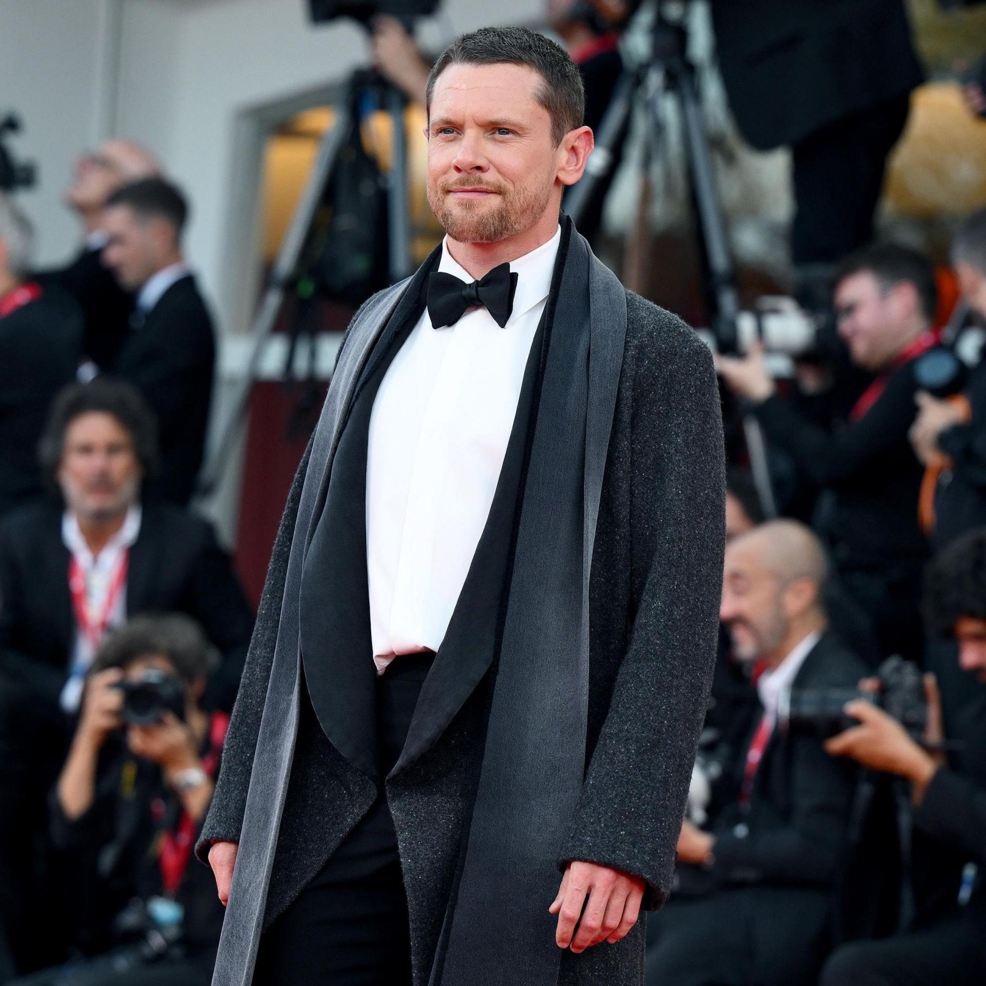 Jack O'Connell looks towards camera while posing on the red carpet wearing a black tie outfit in Italy in September.