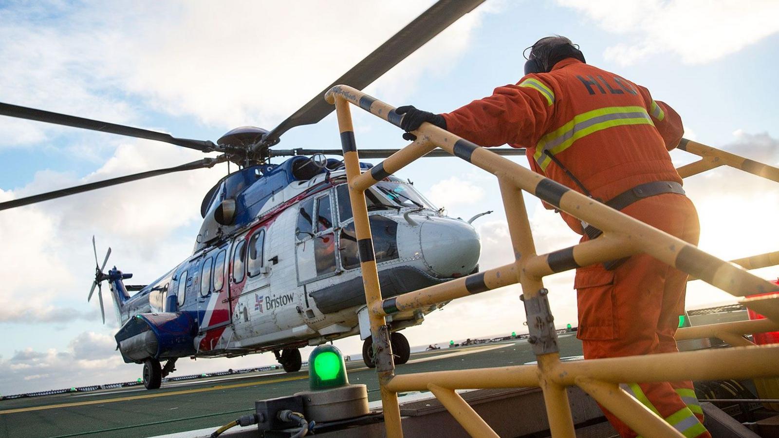 A helicopter lands on an oil rig and a man in orange overalls is climbing up steps and heading towards it
