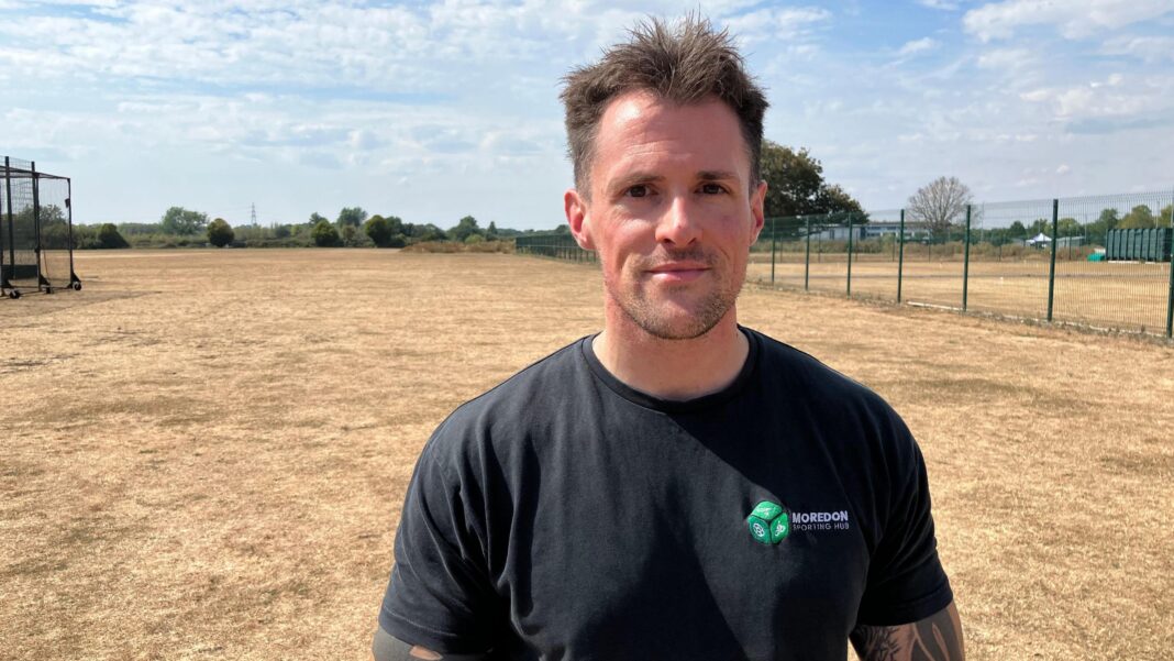Andrew Briggs stands on a dried-out sports pitch. The grass is brown and yellow after weeks of drought. He has short brown hair wears a black t-shirt with a small green logo on the right side of his chest.