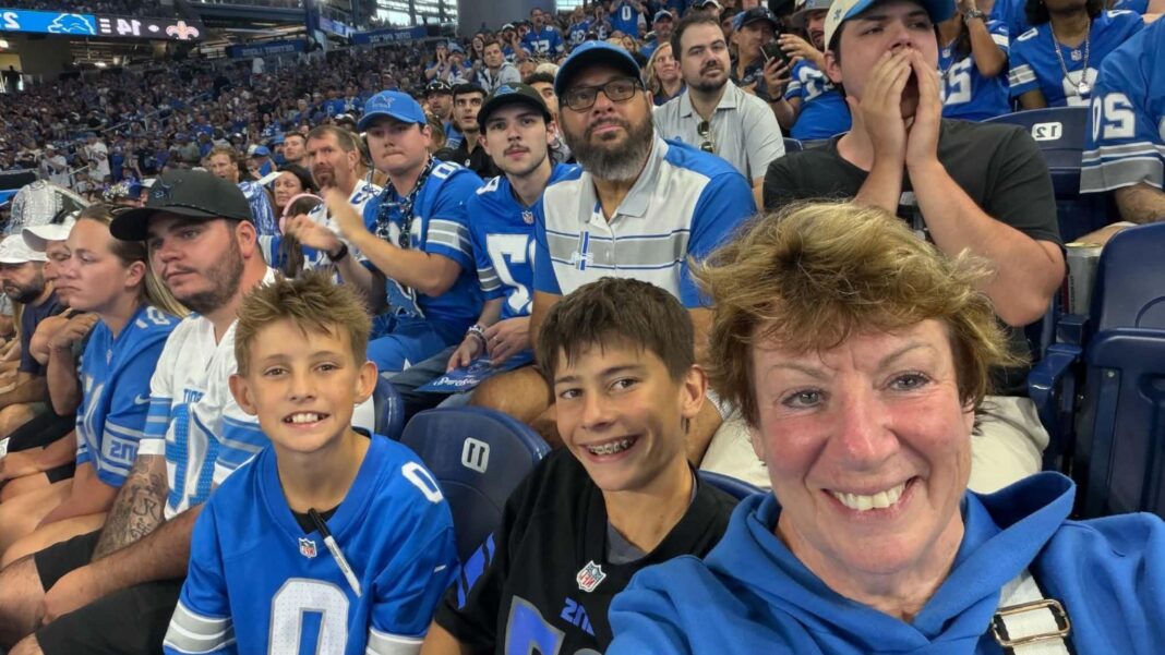 Why an American football game played the Canadian national anthem Carol Breed wearing a blue hoodie takes a selfie at the Lions' stadium with two young boys wearing Detroit Lions. There are other fans in the background.