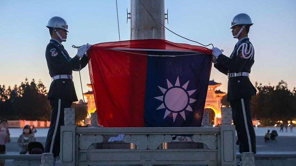 Two ceremonial guards in dark uniforms and white helmets stand on either side of a flagpole, holding a large Republic of China (Taiwan) flag during a flag-lowering ceremony at dusk.