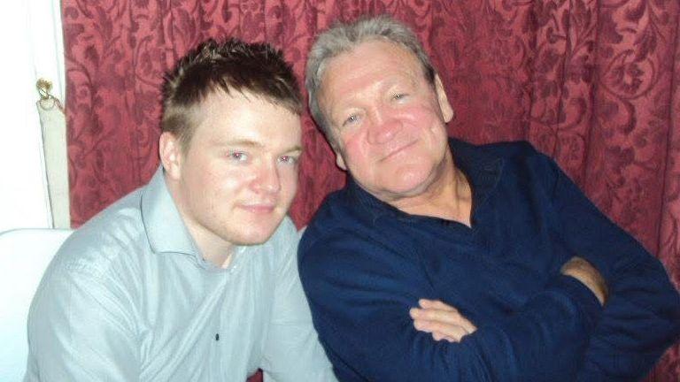 A picture of a father and son. The younger man wears a pale grey shirt and the older man wears a navy shirt with his arms crossed. Both look relaxed and happy.