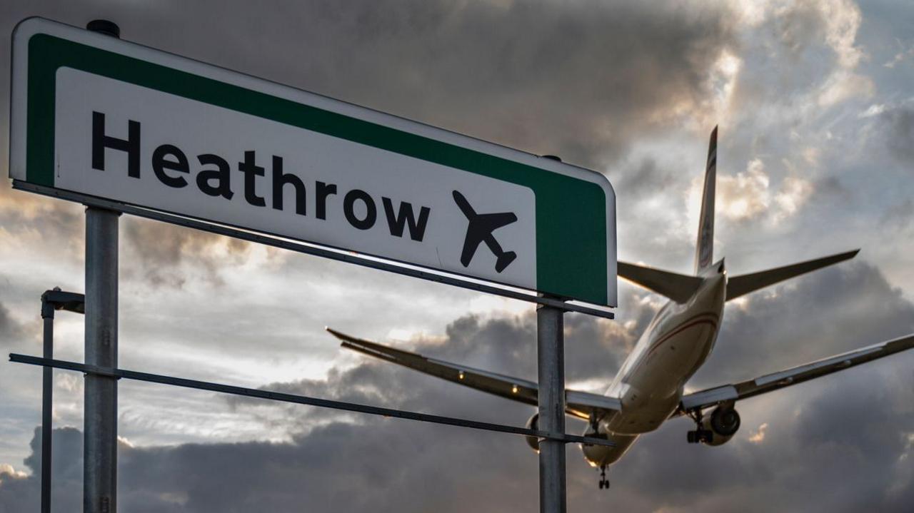 A commercial aeroplane flying low on final approach to land at Heathrow Airport, passing directly over an overhead highway road sign.