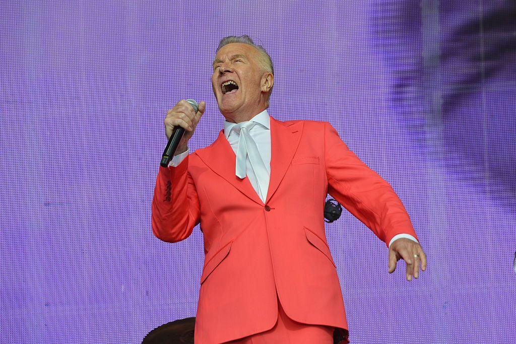ABC's Martin Fry singing into a microphone on stage. He is wearing a red suit, a white shirt and white tie.
