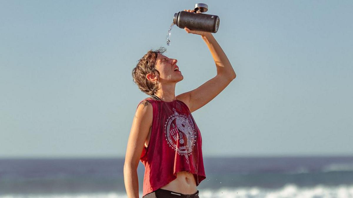 A person is standing on a shingle beach pouring water from a flask onto their head. They are wearing black shorts and a cropped dark red sleeveless top with a white logo. The sky is cloudless and waves can be seen breaking in the background.