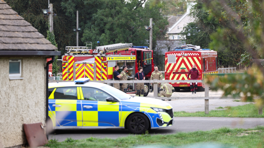 A police car is pictured parked on a road. In the background is a fire engine and crew members