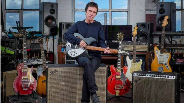 Johnny Marr is sitting on a large amp holding a black and white coloured guitar. Next to him is another amp and behind him are five guitars and other musical equipment