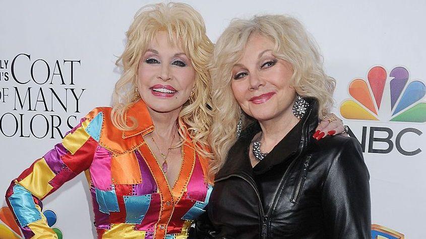 Dolly Parton, wearing a bright and colourful dress, stands next to her sister Stella at a red carpet event in 2015 - who is wearing a black leather jacket and large jewellery. They are both smiling, with Dolly's arm around her sister's shoulders, as they are photographed.