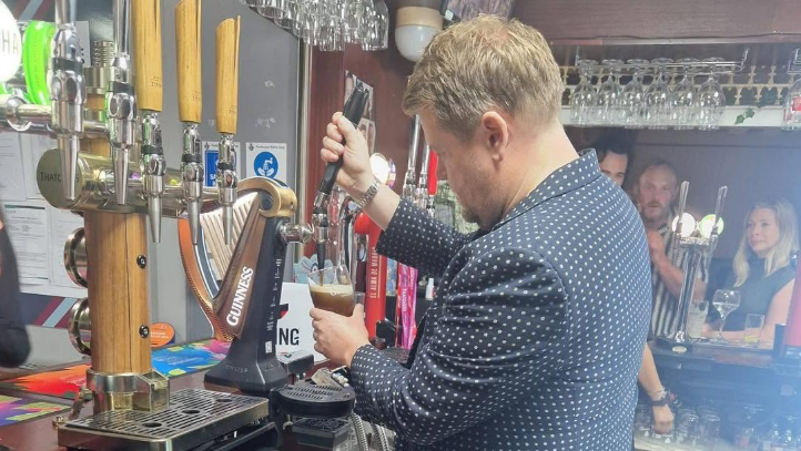 James Corden pouring a pint of Guinness behind the bar as customers see him doing it.