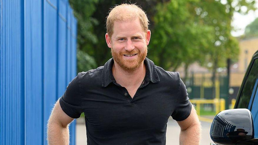 Prince Harry wearing a black polo shirt smiles at the camera.