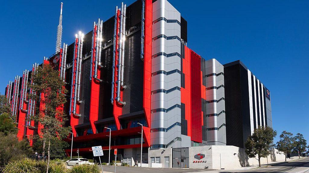 A huge red and grey windowless building with pipes on the outside against a blue sky.