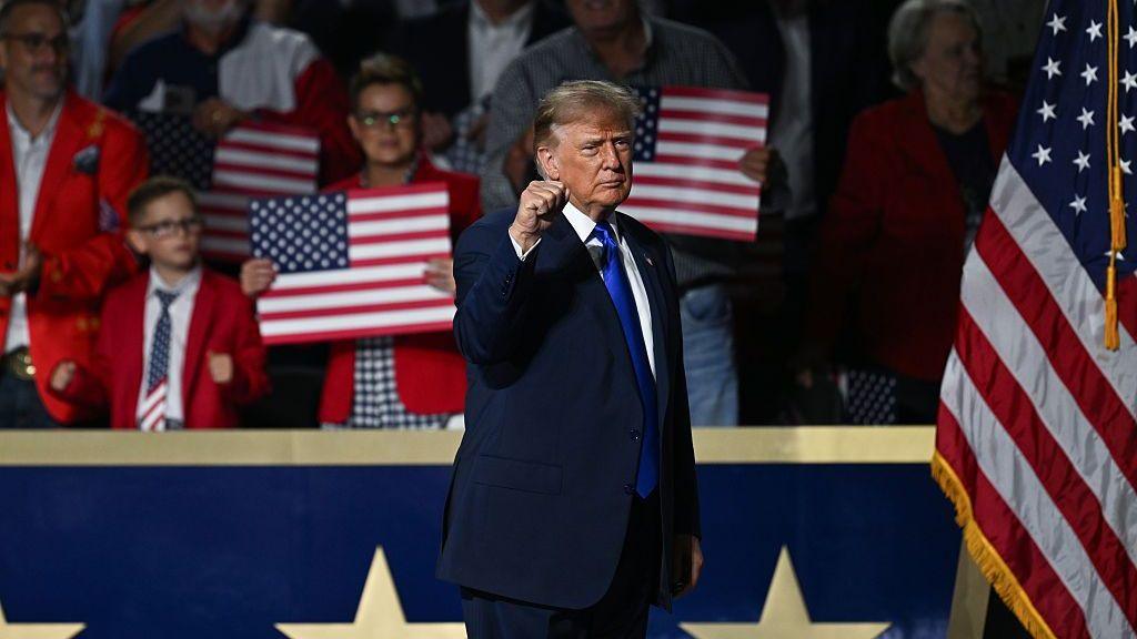 President Trump, in a blue suit and tie" raises a fist on stage. Behind him, people are gathered and holding American flag signs.