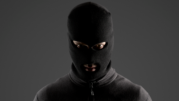A stock image of a man wearing a black hoody and black balaclava standing in front of her black wall.