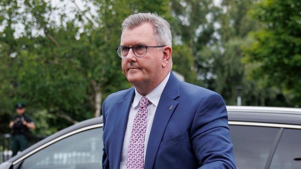 Jeffrey Donaldson is walking away from a black car. He is a man with short grey hair, wearing a dark blue suit, white shirt and pink tie.