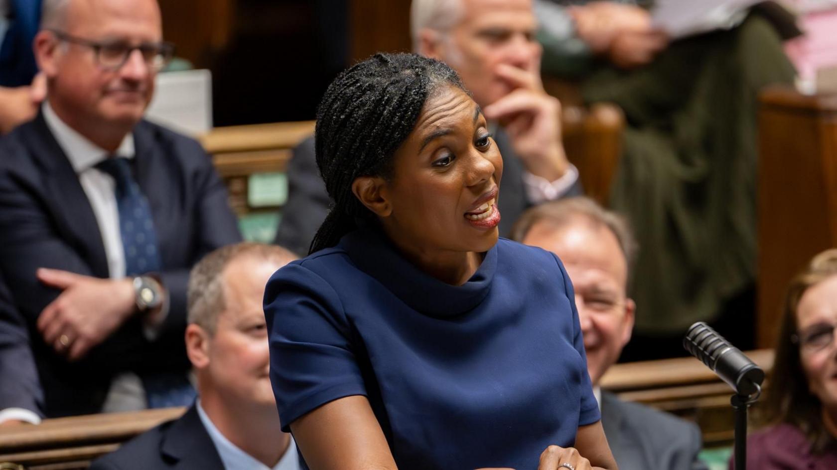 Kemi Badenoch speaking in the House of Commons at PMQs. She is wearing a blue dress with a high collar