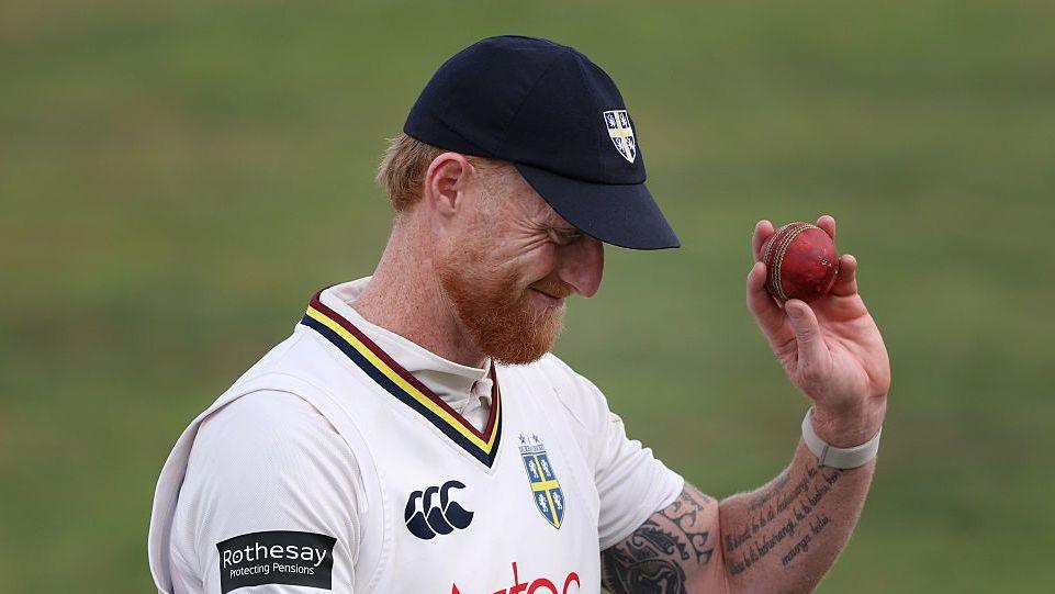 Ben Stokes raises the red ball in his left hand as he bows his head, while wearing his navy Durham cap