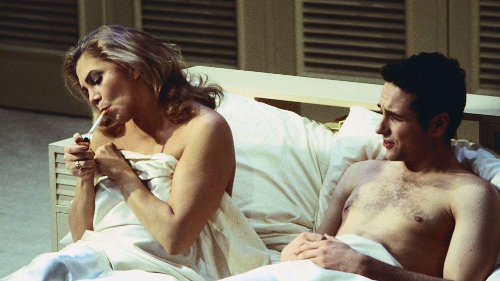 A woman and man in bed on stage. She is lighting a cigarette