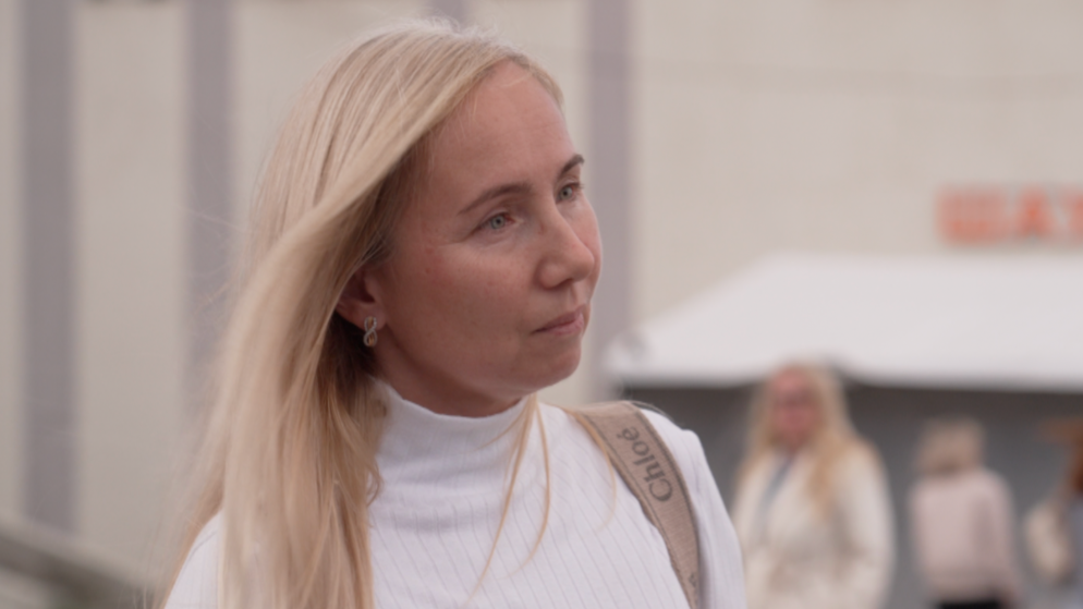 Olga stands in a white top with a Chloé bag strap visibly slung over her shoulder. She has blonde hair and earrings in the shape of a number 8.