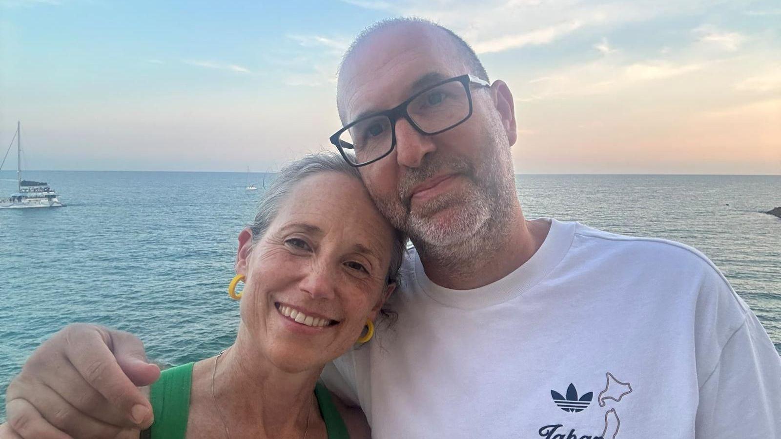 Kate rests her head on her husband's shoulder while he puts his arm around her. They both smile for the camera against a backdrop of the sea.