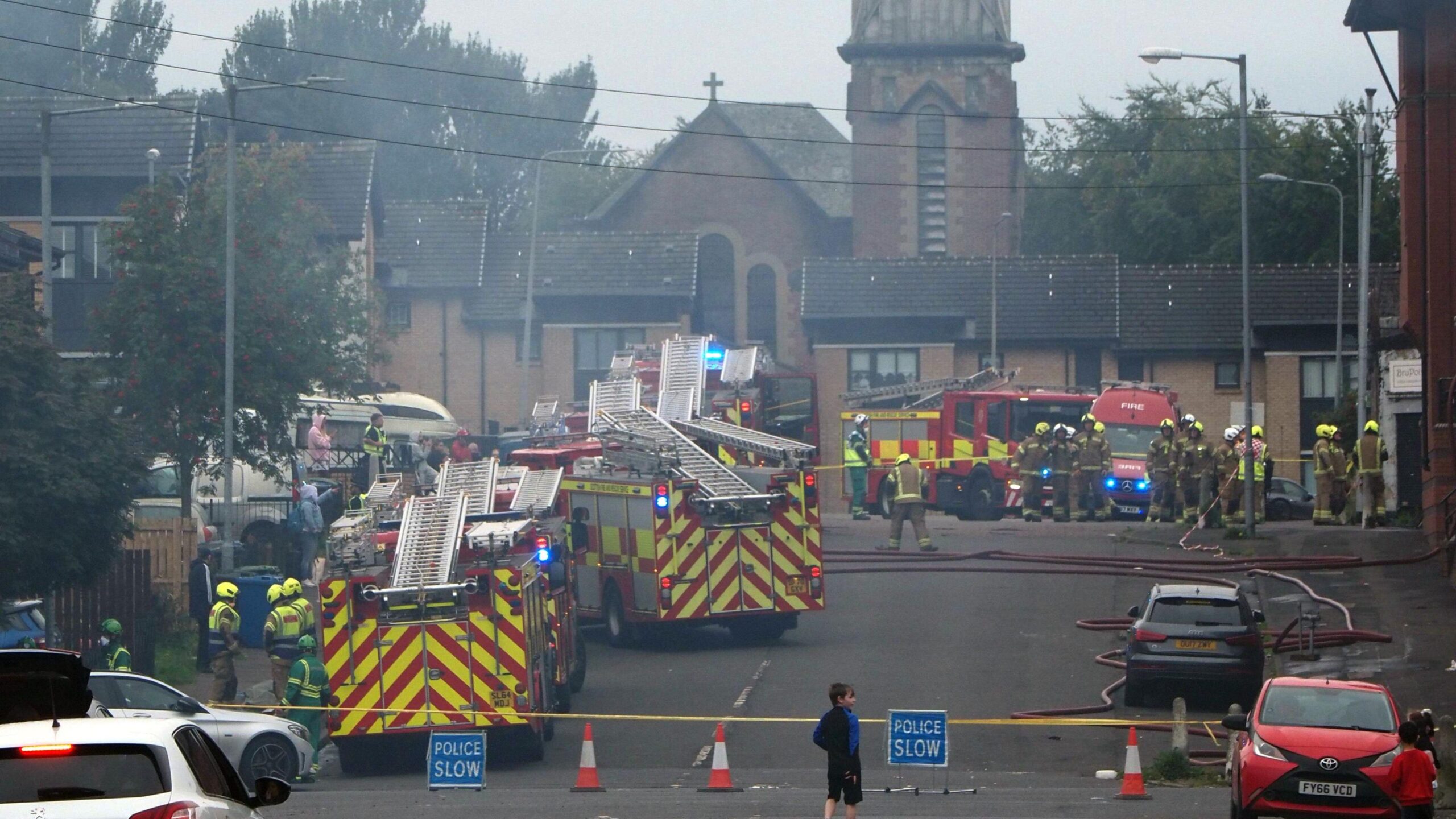 Four fire vehicles and more than a dozen firefighters in an area of a street which has been cordoned off with police tape and signs. There is a church with a steeple at the end of the road and parked cars.