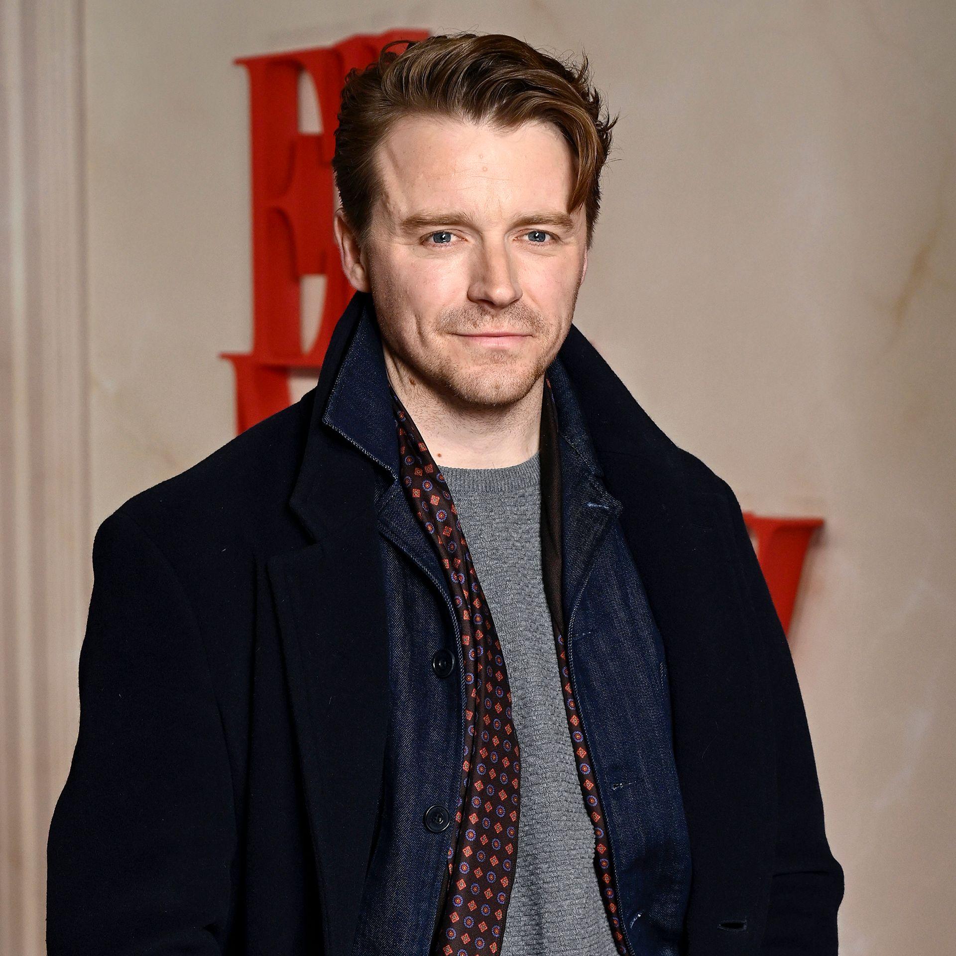 Jack Lowden looks at camera while posing for a photo in a black coat and grey jumper, in London in 2025.