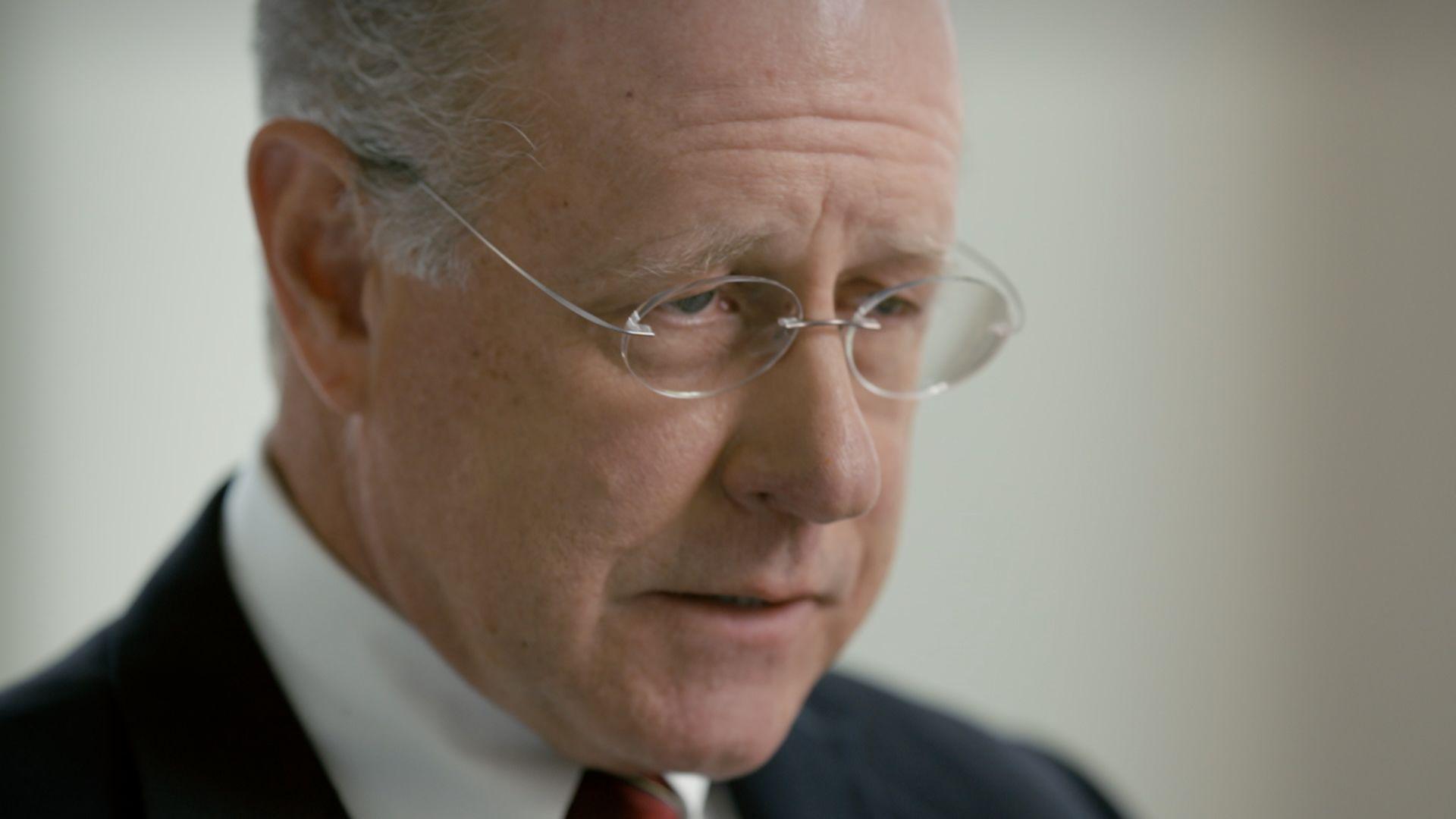 Rick Lambert, a man with grey hair at the side of his head and oval, rimless glasses, pictured in close-up with shallow depth of field, wearing a dark suit with a tie, looking downwards with a blank wall behind him