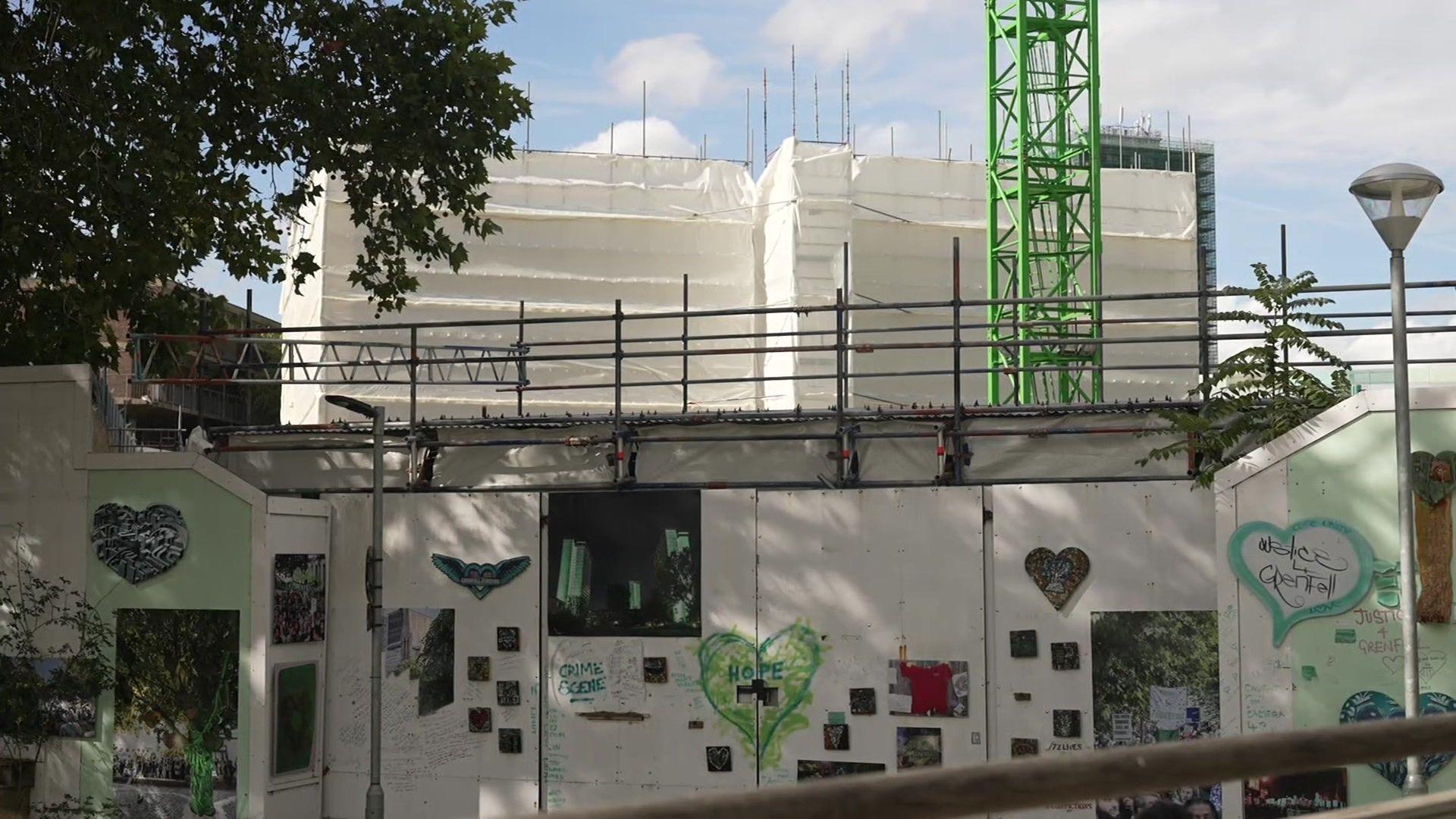 Grenfell Tower covered in protective white sheeting during dismantling work, with scaffolding and a green crane above a memorial wall decorated with hearts, photographs and tributes.