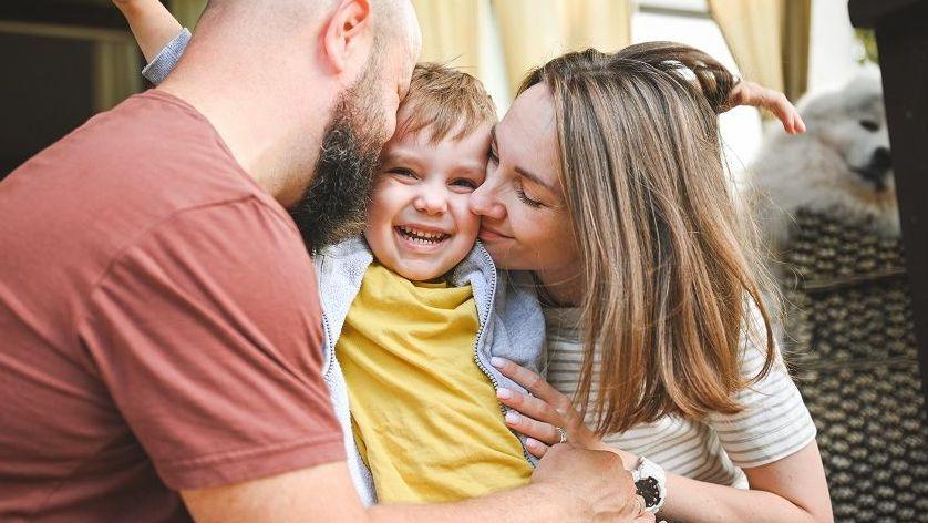 A young blonde child wearing a yellow t-shirt is hugged by a man wearing a red t-shirt and a woman wearing a beige striped t-shirt.