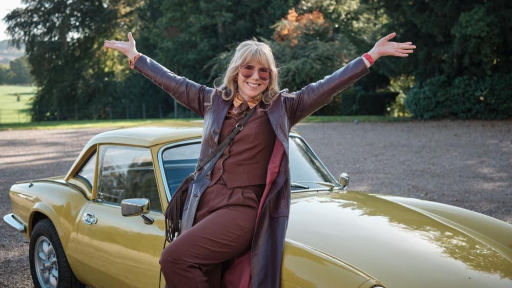 A woman with long blonde hair and large sunglasses poses with her arms in the air in front of a mustard coloured, two seater car. She's dressed in a brown two piece with a long brown leather coat over the top. The car is parked on gravel with trees and countryside in the background.