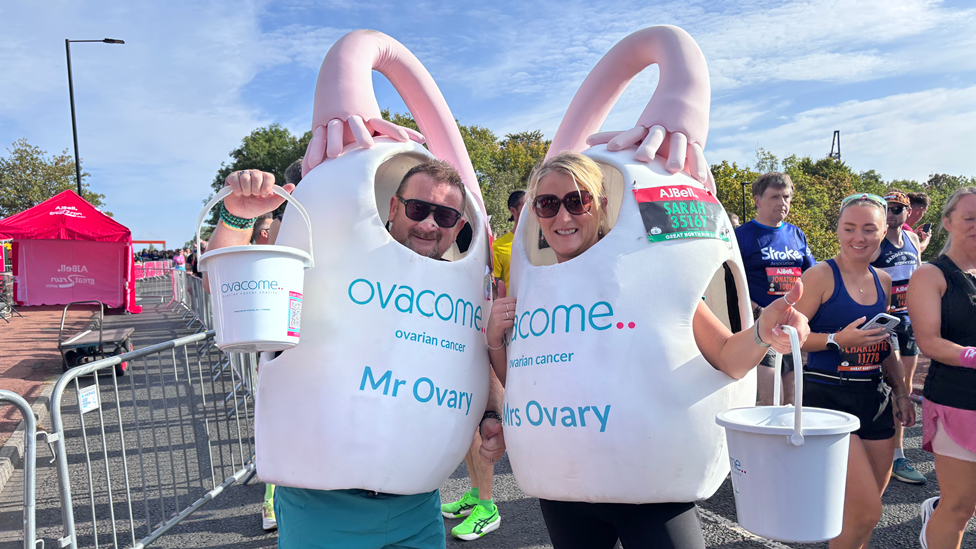 Two race participants dressed as giant ovaries hold fundraising buckets for the ovarian cancer charity Ovacome. It is a white sphere with a pink design coming out the top.