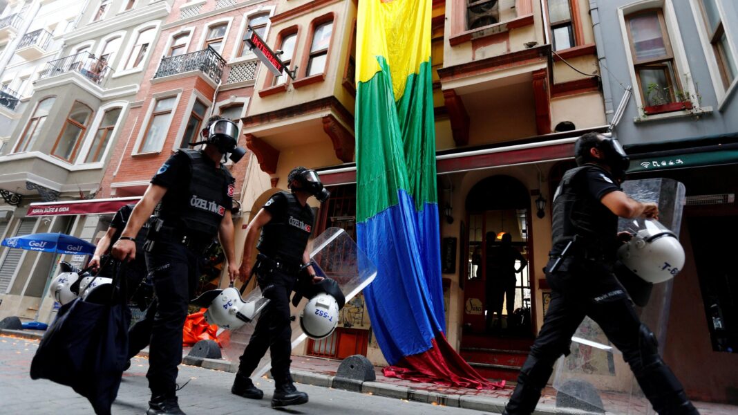 Riot police, with a rainbow flag in the background, chase LGBT activists as they try to gather for a pride parade, which was banned by the governorship in Istanbul. Officers are wearing black uniforms, wearing gas masks, and are carrying helmets and riot shields as they move through what appears to be a residential street.