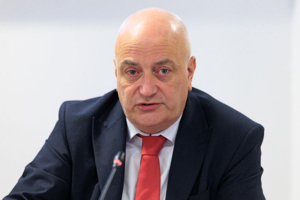A bald man in a navy suit, blue shirt and red tie
