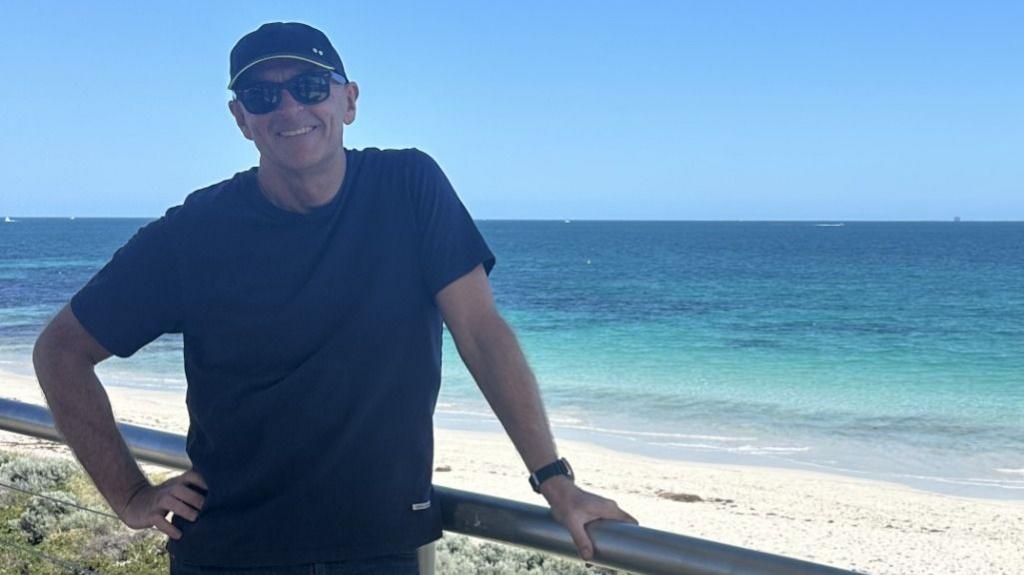 Greg O'Neill stands beside a beachfront railing overlooking clear blue water and white sand on the Western Australian coast.